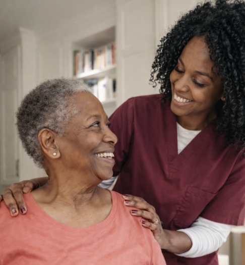 African American caregiver smiles and rests her hands on an older woman’s shoulders as they share a warm moment in a bright home.