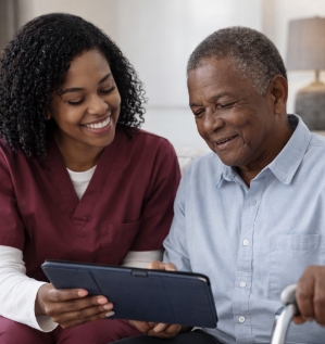 Caregiver and older man sit together on a couch, smiling as they review information on a tablet.