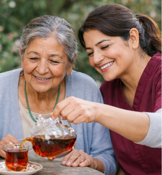 Caregiver pours tea into a small glass cup for a smiling older woman at an outdoor table in a garden.